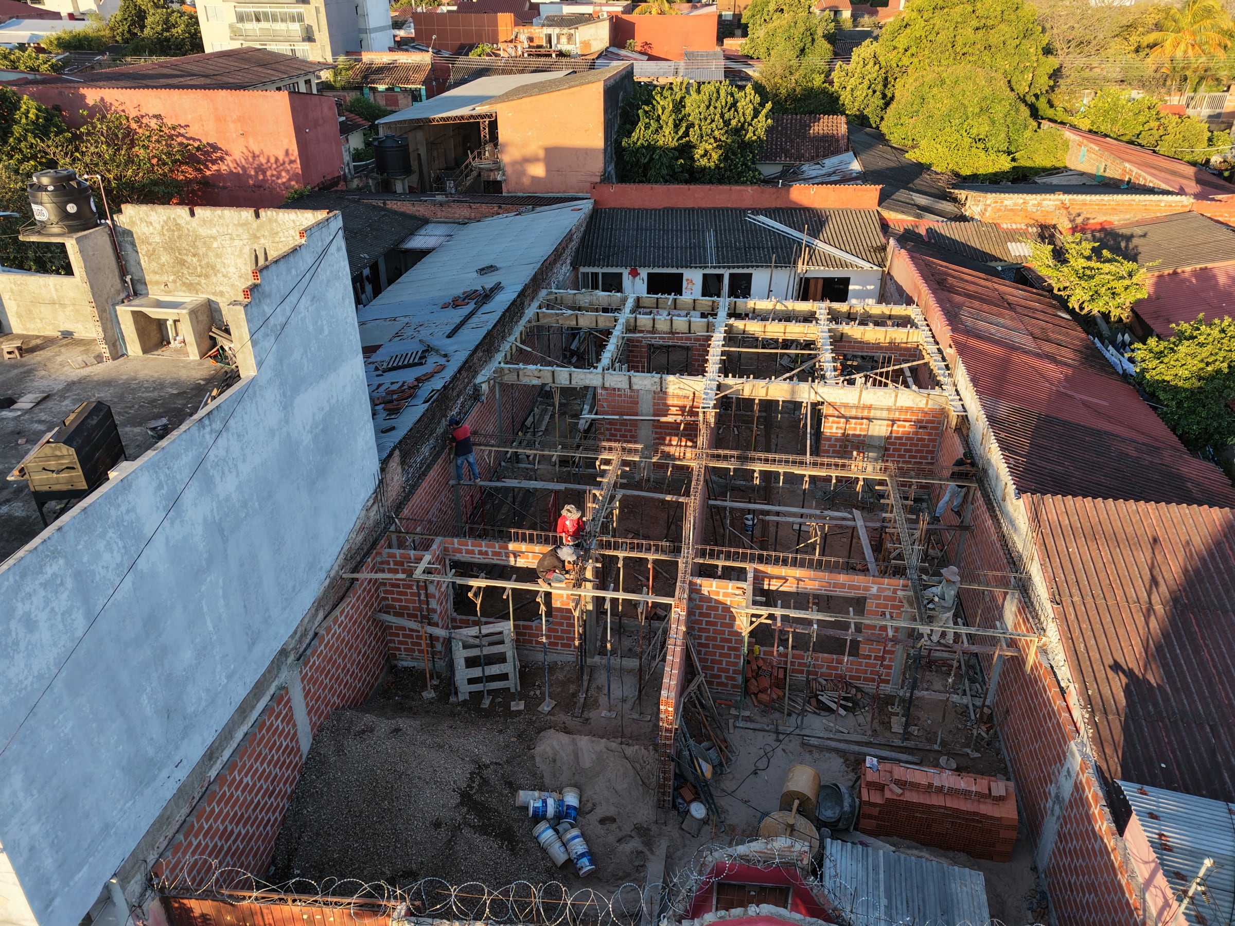 Vista aérea de la obra de la calle 8: muros de ladrillo levantados, columnas de hormigón armadas y encofrado de la losa, con obreros trabajando.